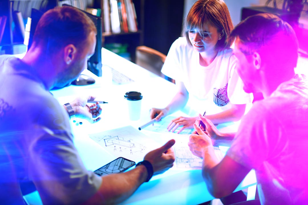 Three people working together at a table with digital devices in a dimly lit room.